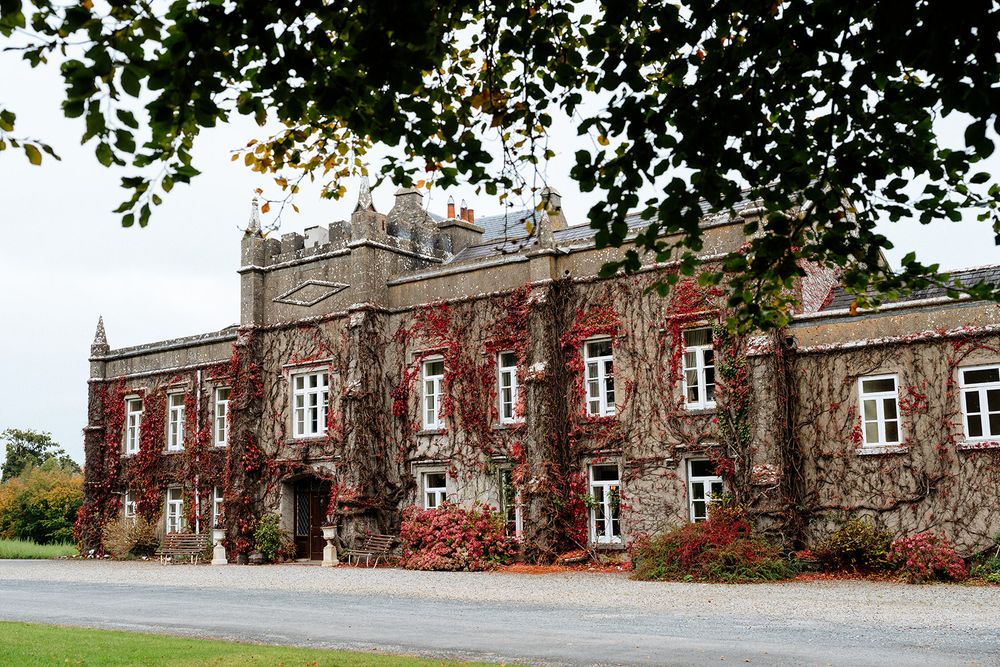 Historic stone building with red ivy-covered walls and Gothic architectural details stands on autumn grounds.