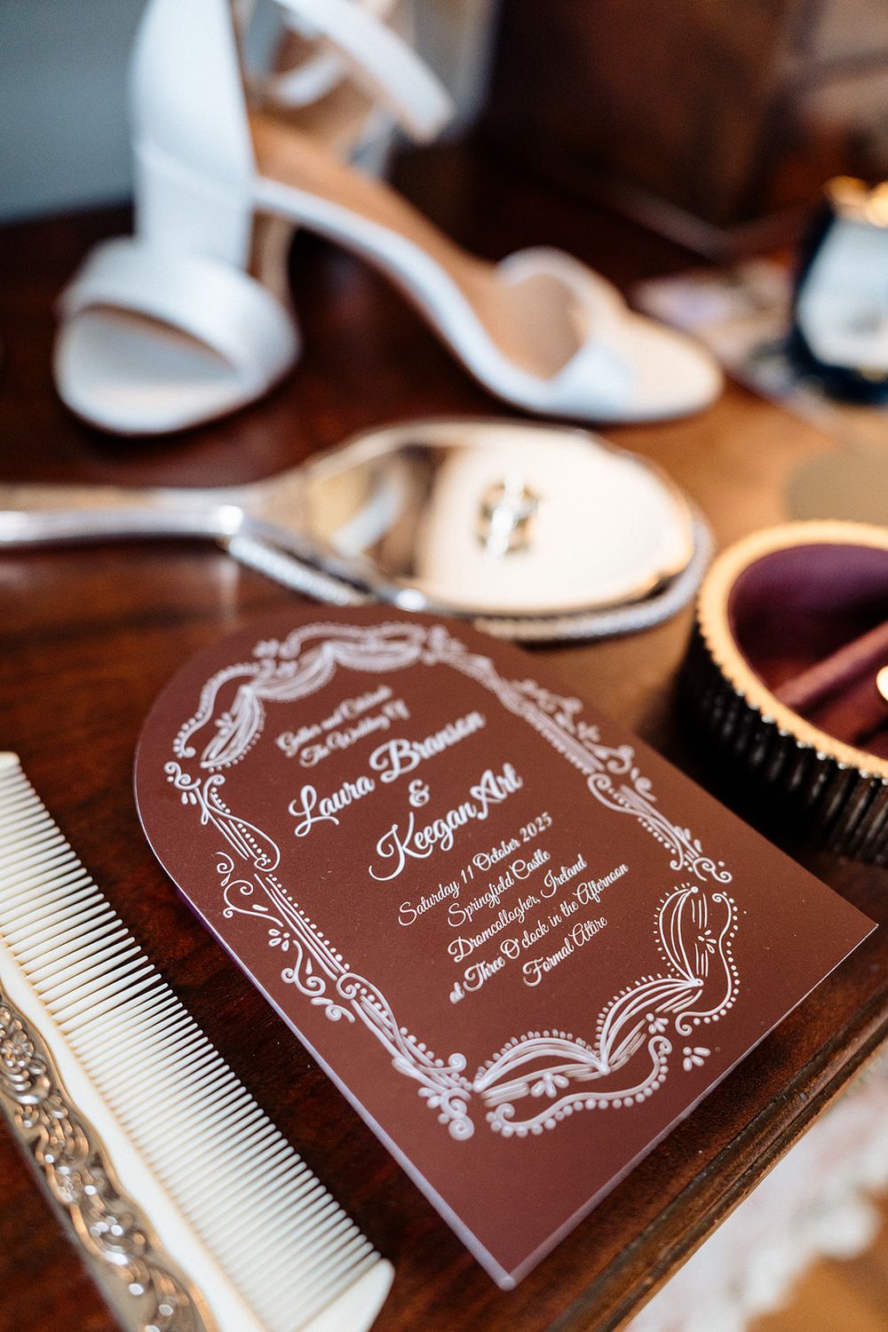Elegant brown wedding invitation cards with white ornate designs displayed alongside white decorative combs on a wooden surface.
