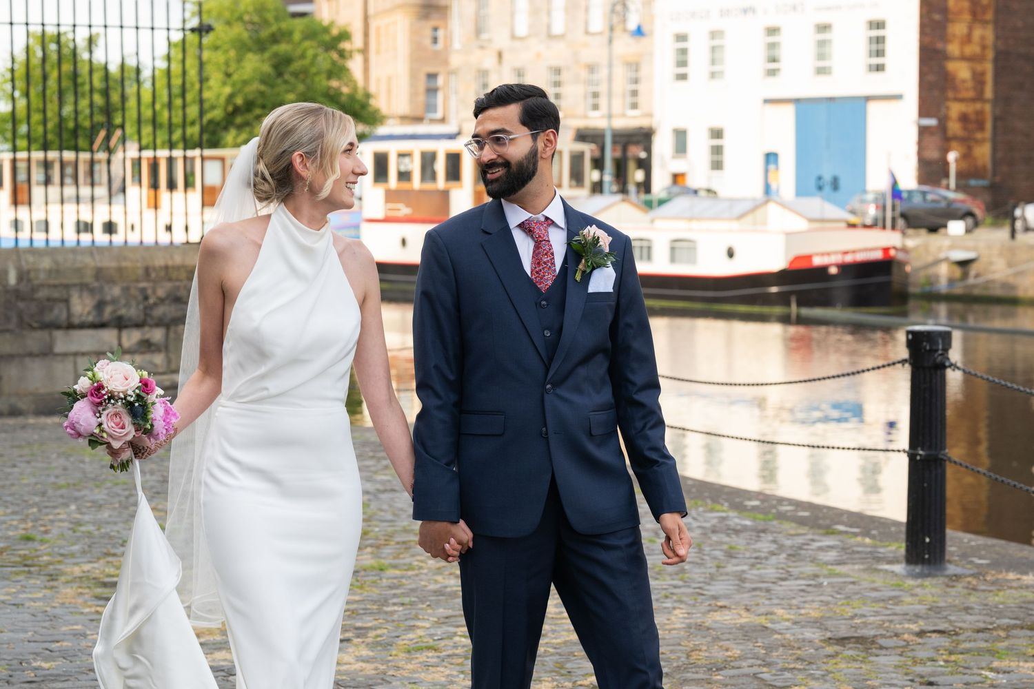 Bride and groom walking hand in hand along waterfront path.