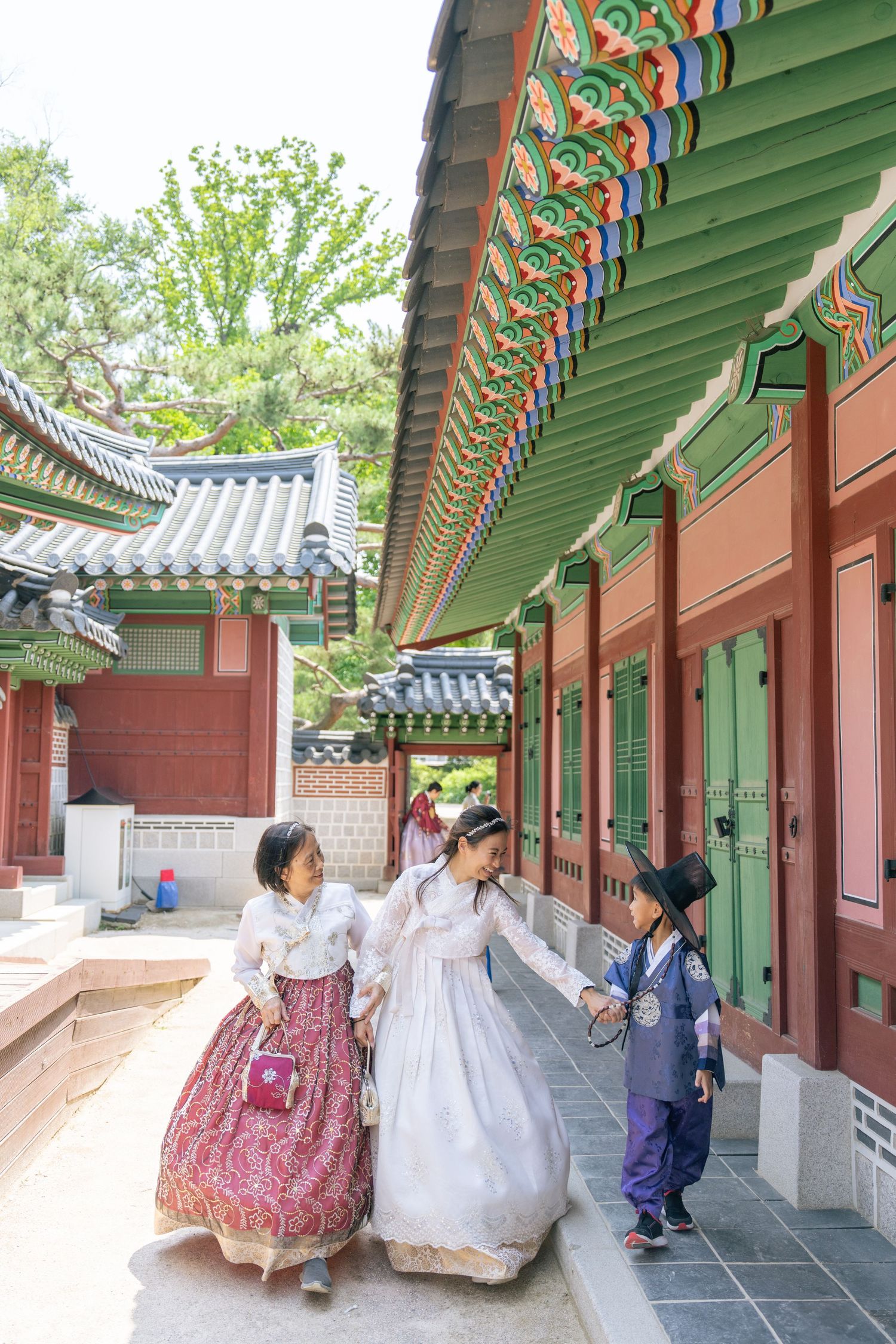 People in traditional Korean hanbok clothing walk along a covered outdoor corridor with ornate painted wooden beams and green roof tiles.
