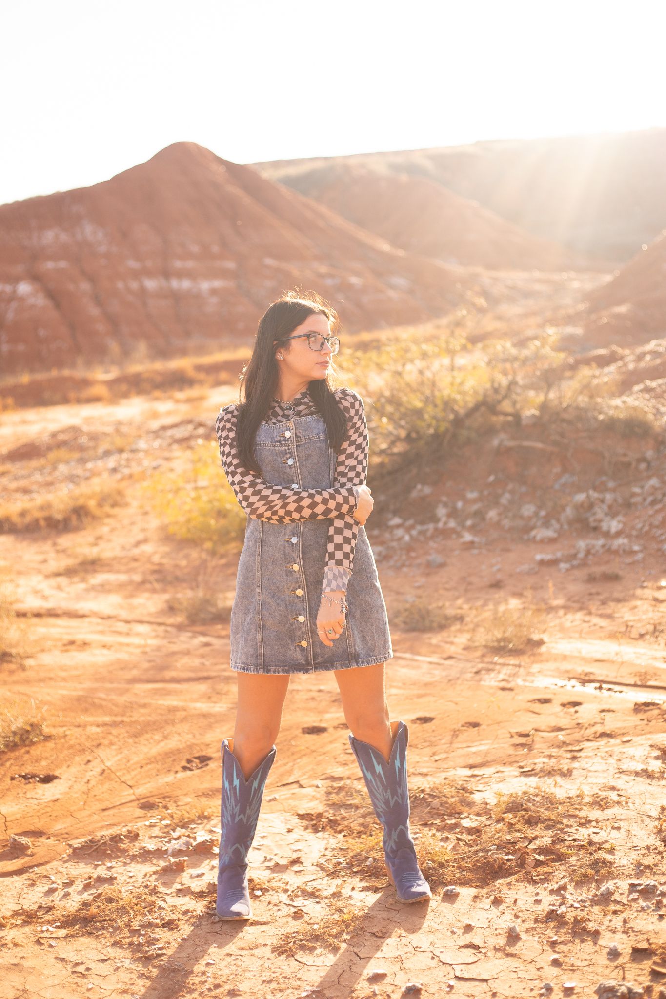 A person in denim and checkered top poses in golden sunlight on a rocky desert landscape.