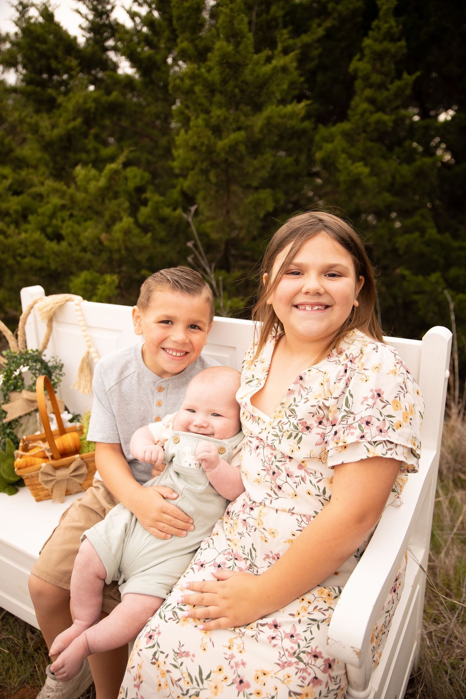 A family sits together outdoors on white chairs against a backdrop of evergreen trees in warm sunlight.