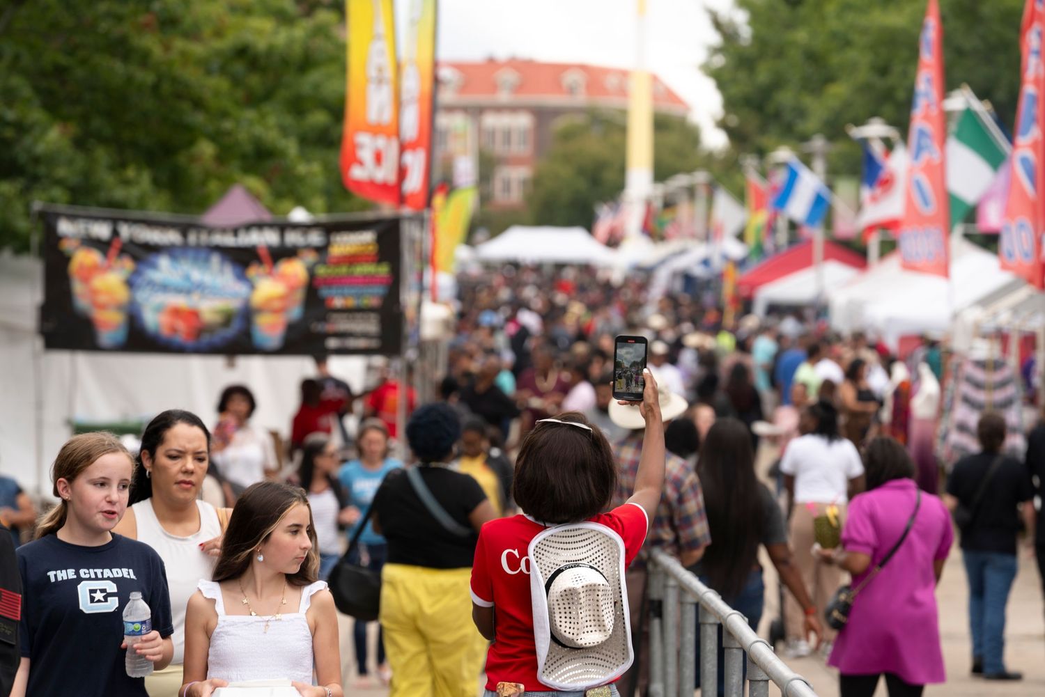 Crowds of people walking through a busy outdoor festival with vendor tents and flags lining the street.