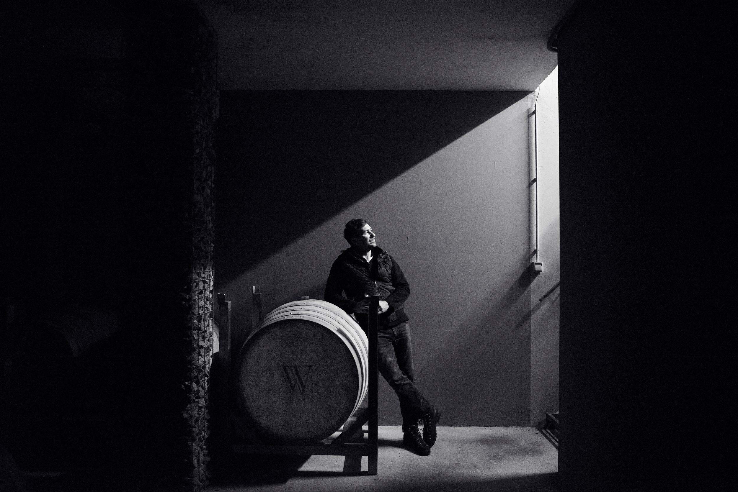 Dramatic black and white photo of a winemaker leaning on a barrel in a beam of light.