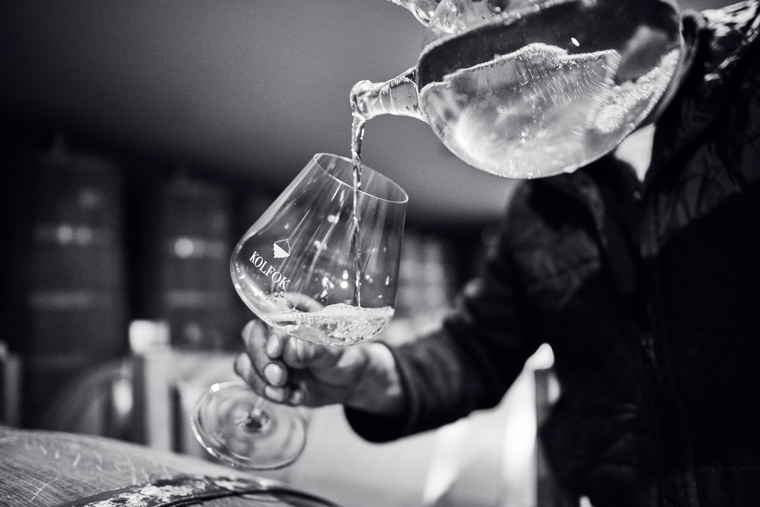 Black and white close-up of wine being poured from a decanter into a branded tasting glass.