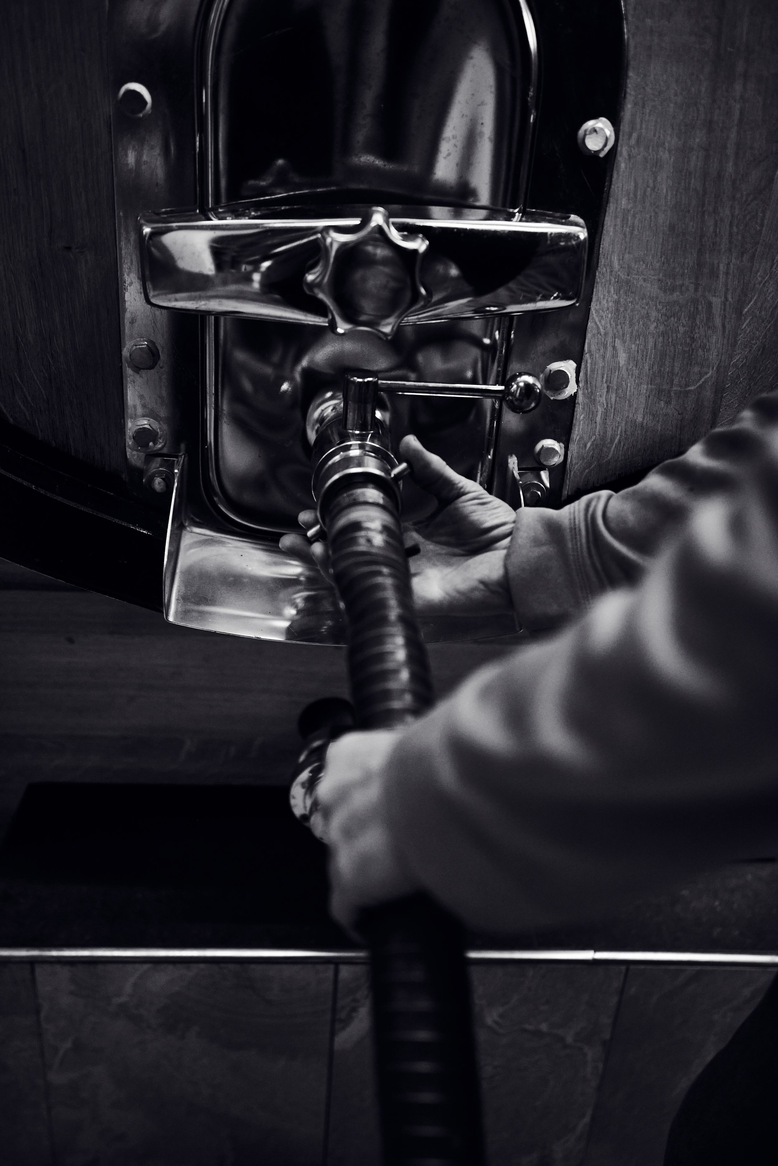 Black and white close-up of hands connecting a hose to a wine barrel valve in a cellar.