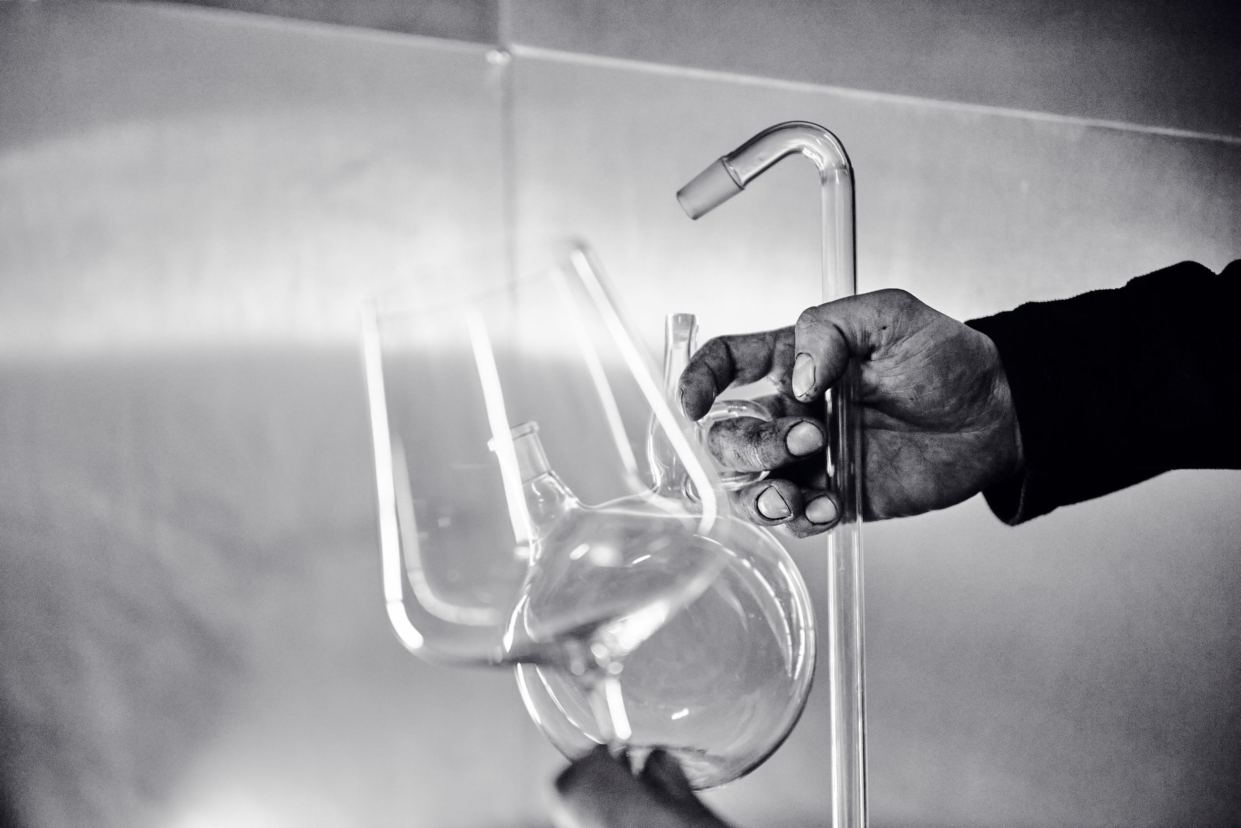 Black and white close-up of hands handling glass laboratory distillation equipment in a winery.