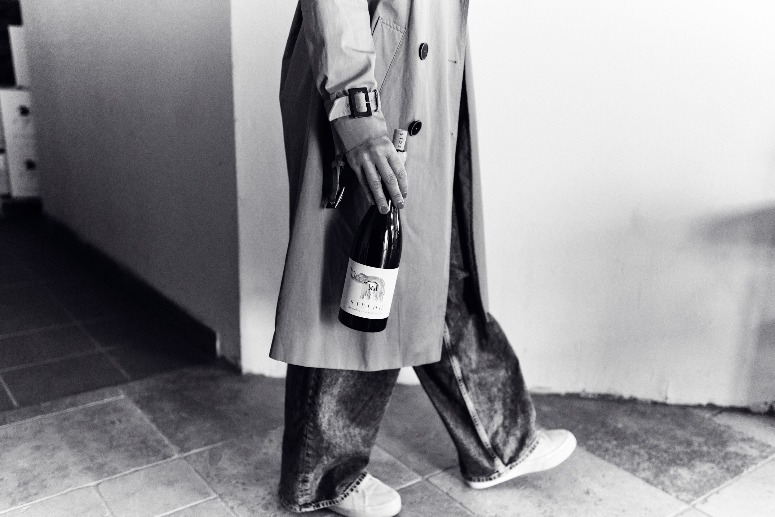 Person in trench coat carrying a wine bottle while walking on tiled floor, black and white photography.