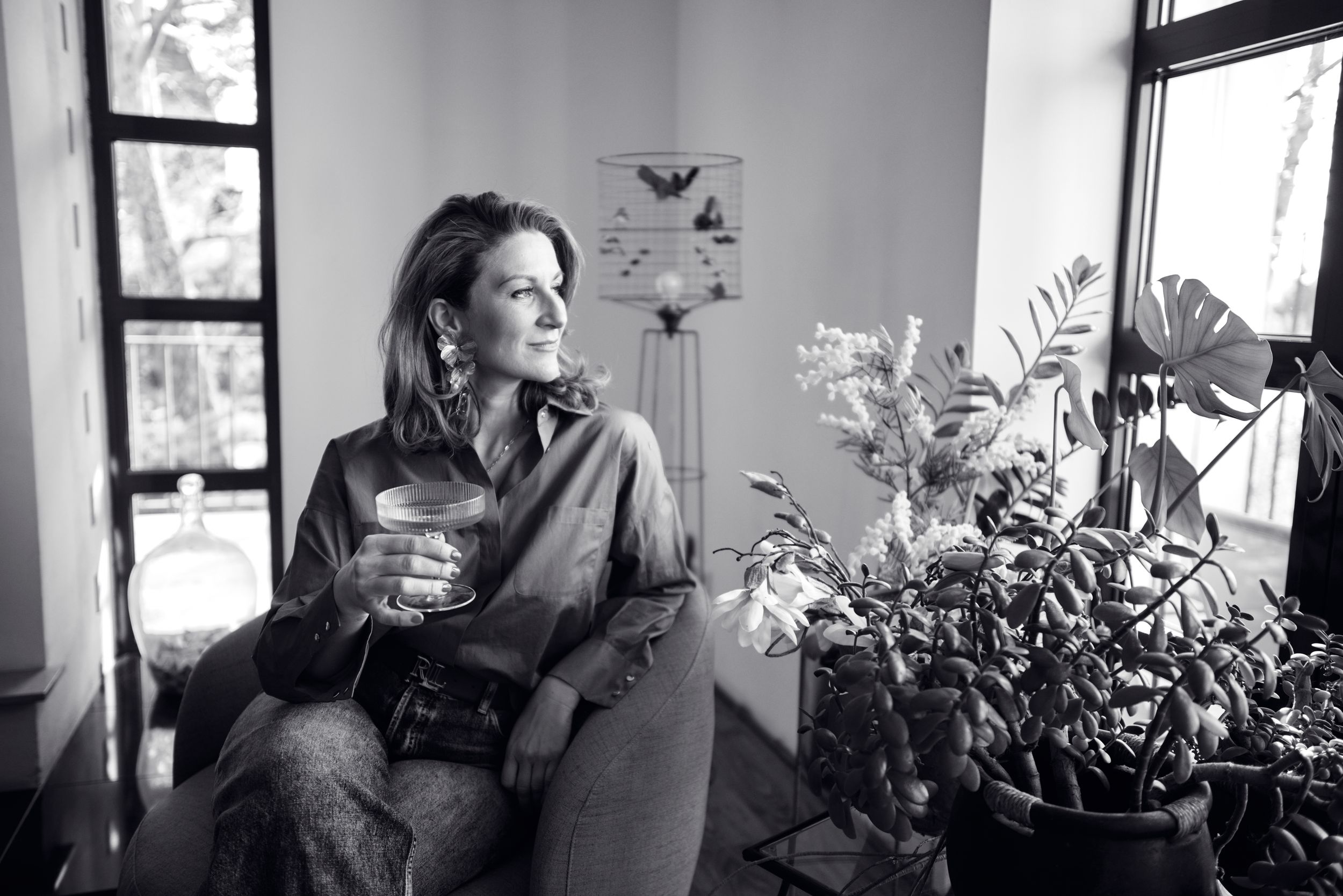Woman sitting by window holding a wine glass surrounded by indoor plants, black and white portrait.