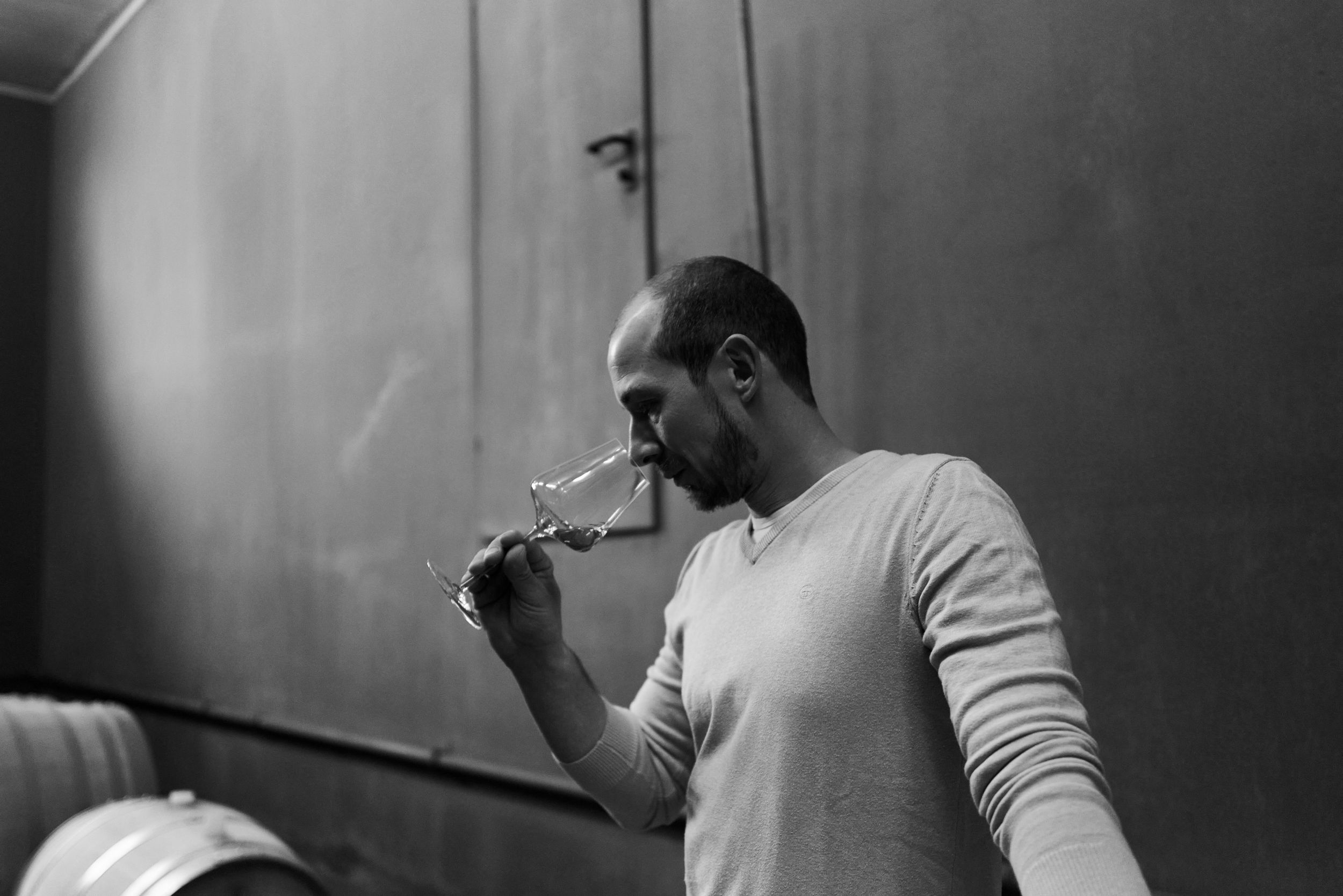 Man in white sweater tasting wine from a glass in a dimly lit cellar, black and white photo.