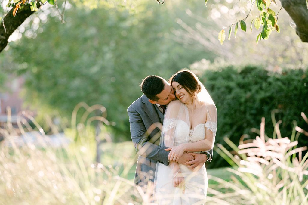 A couple in formal attire shares an intimate moment while walking through a sunlit park setting.