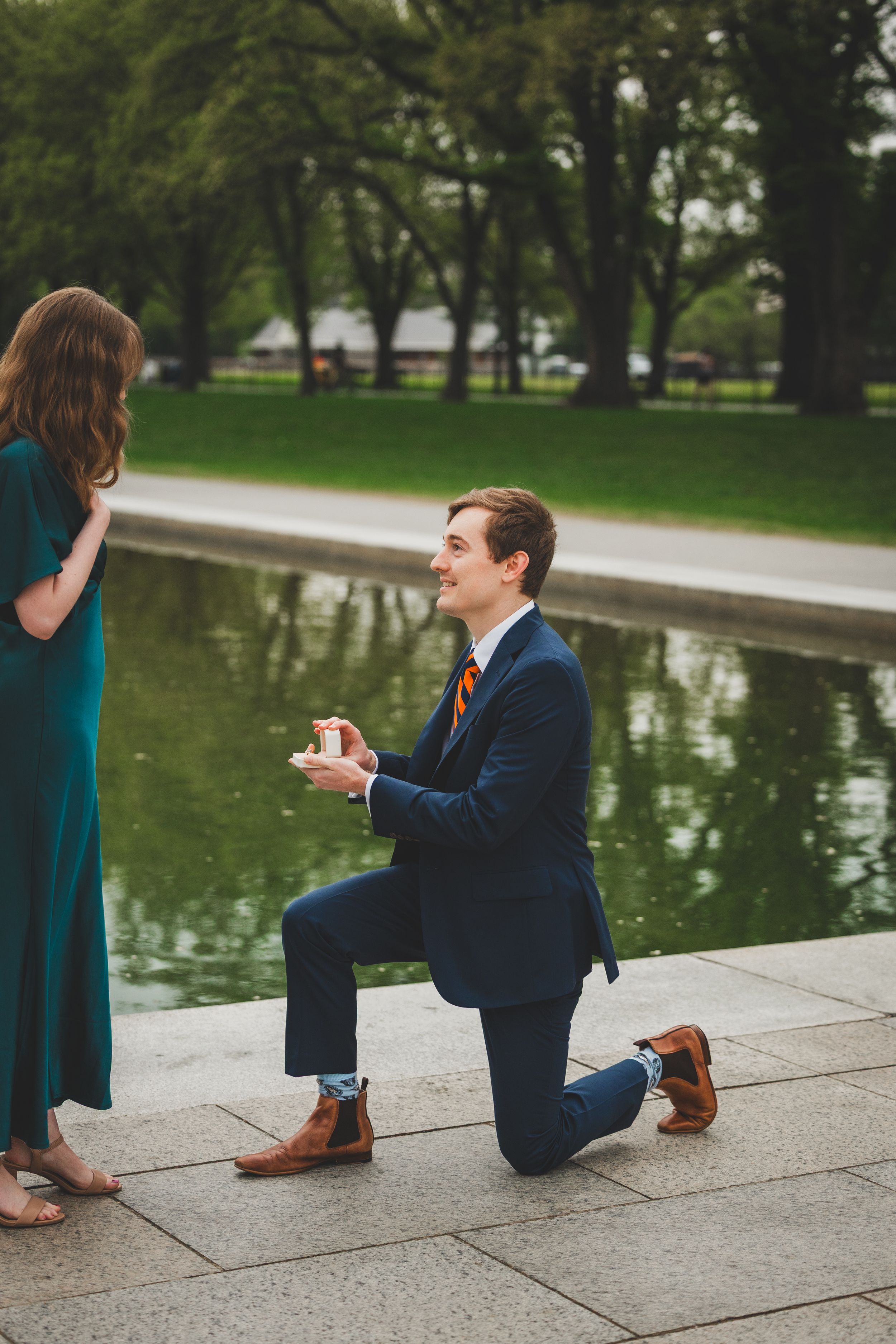 A romantic proposal sequence captured by a lake showing someone in a navy suit kneeling on stone steps.