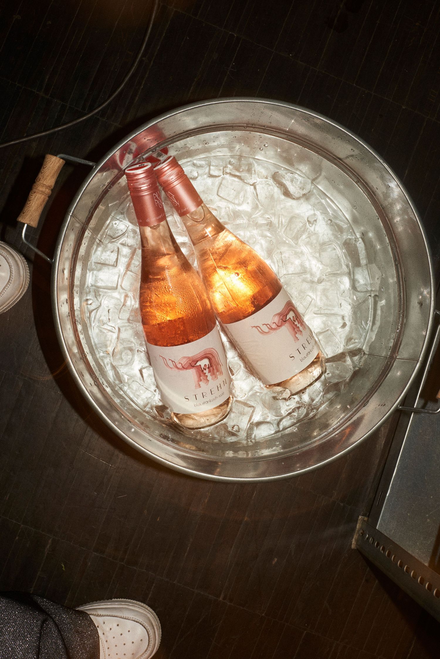 Two rose wine bottles chilling in ice-filled silver bucket viewed from above on dark floor.