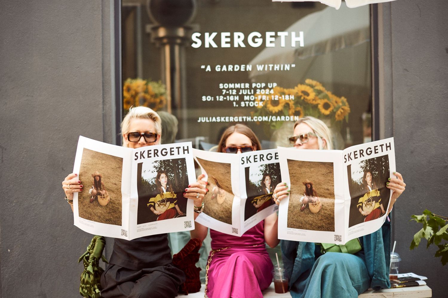 Three women sitting outdoors holding Skergeth A Garden Within summer pop-up newspapers.