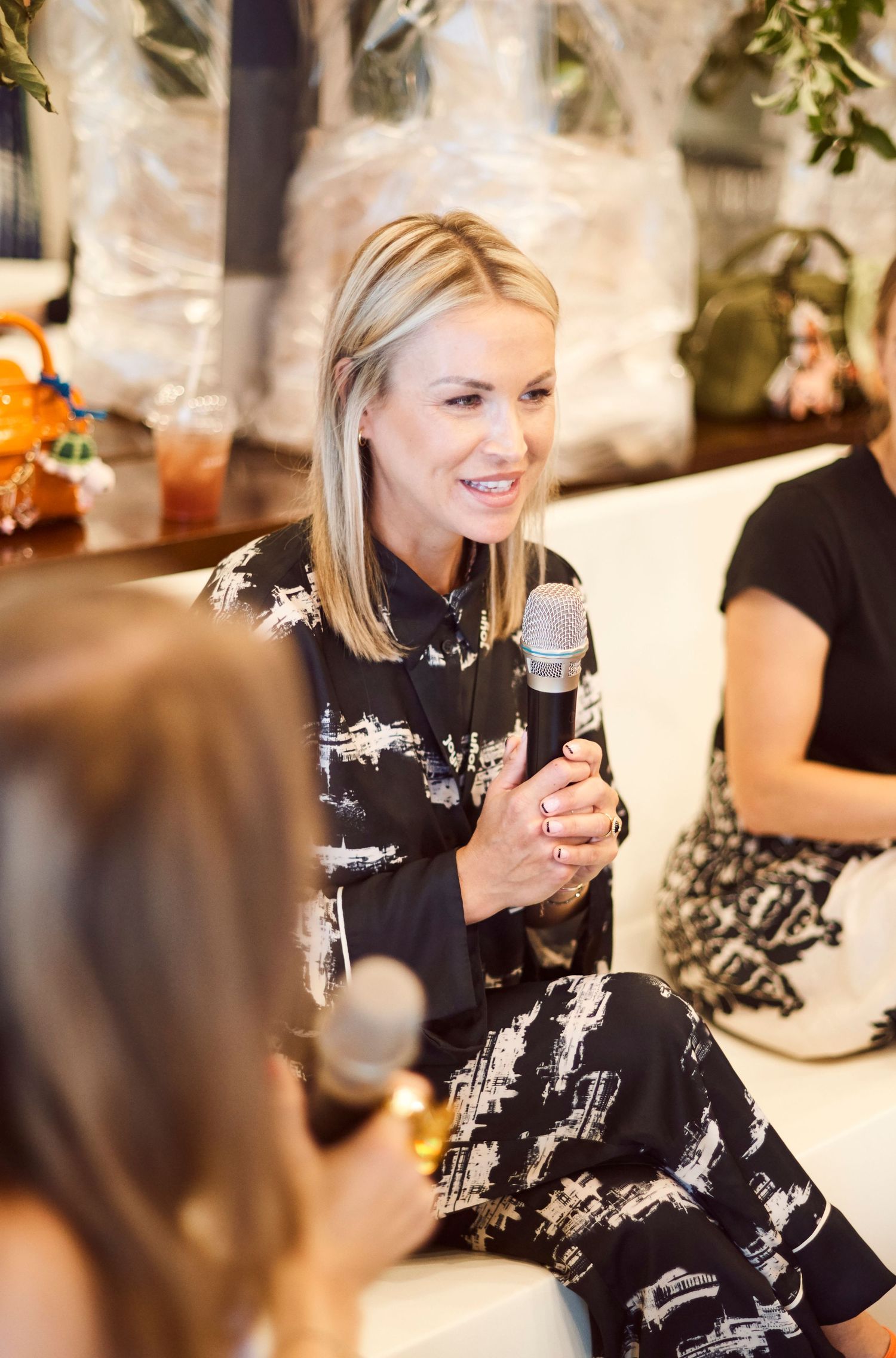 Elegant blonde woman in patterned dress speaking into microphone at indoor panel event.