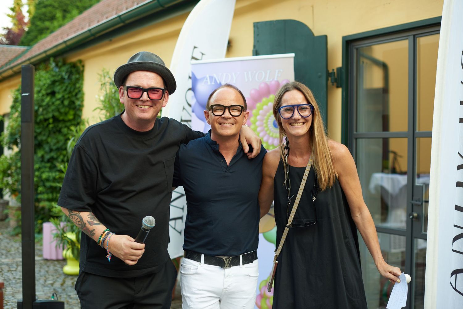 Three people posing outdoors at a summer event, man holding microphone, wearing stylish casual outfits.