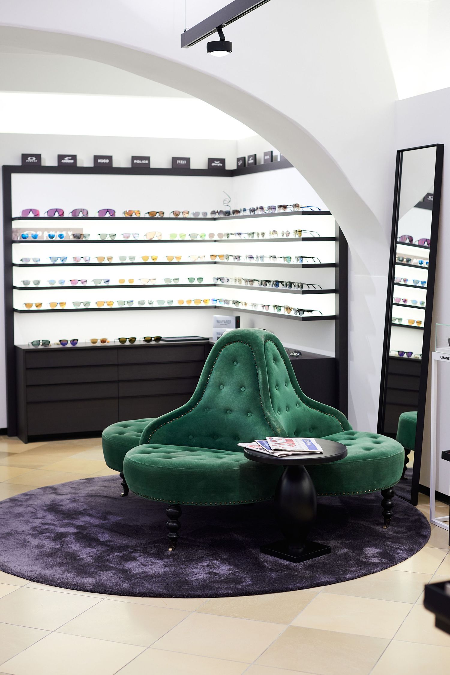 Elegant green velvet tufted sofa in a modern eyewear boutique with sunglasses displayed on black shelving.