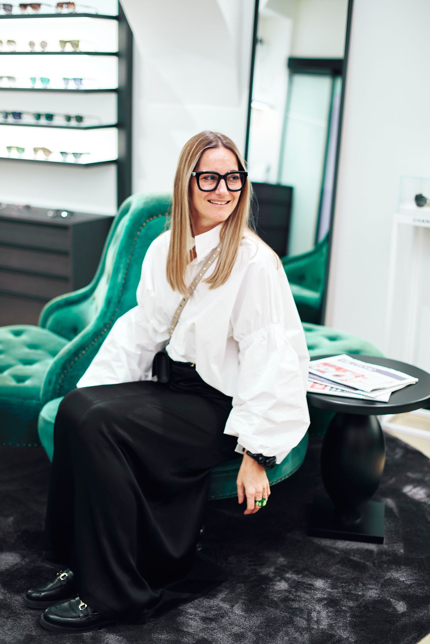 Smiling blonde woman wearing black-framed glasses and white blouse seated in green velvet chair in eyewear store.