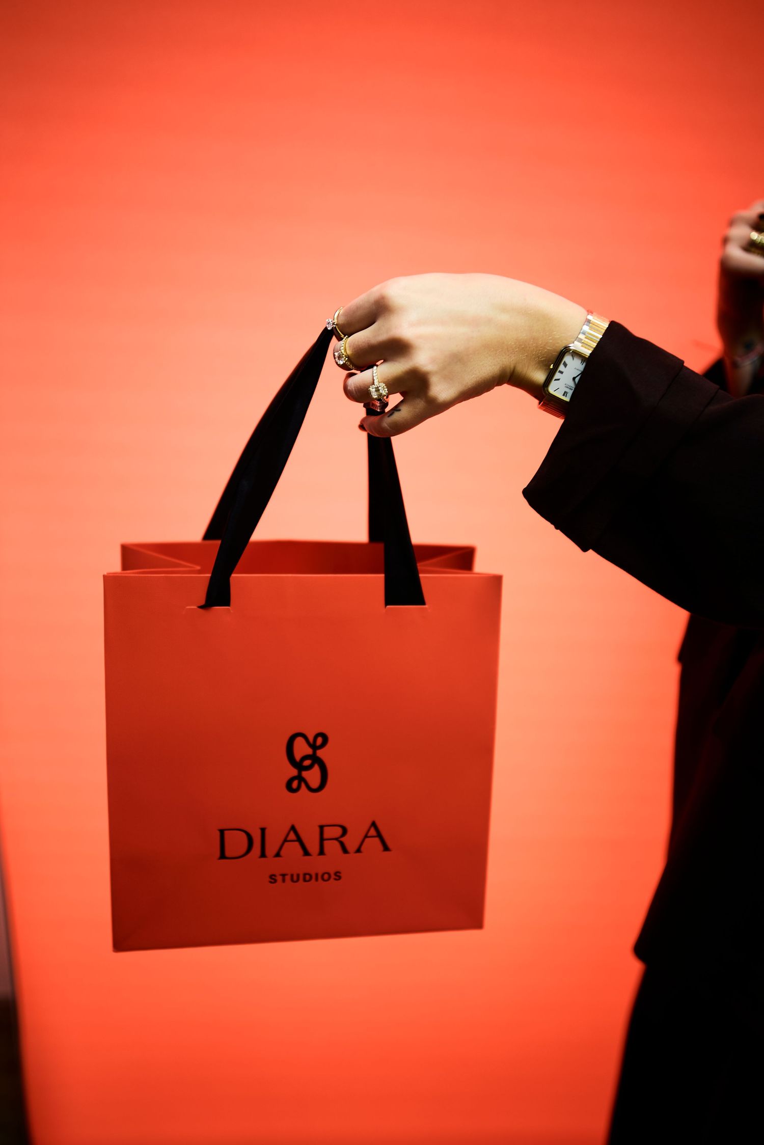 Hand holding red Diara Studios shopping bag with black ribbon handles on red background.