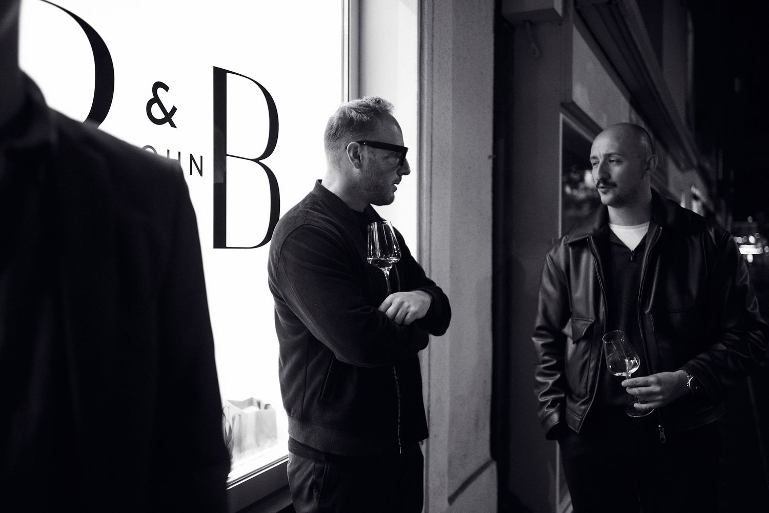Two men in black outfits conversing in front of a D&B store window in a black and white photo.