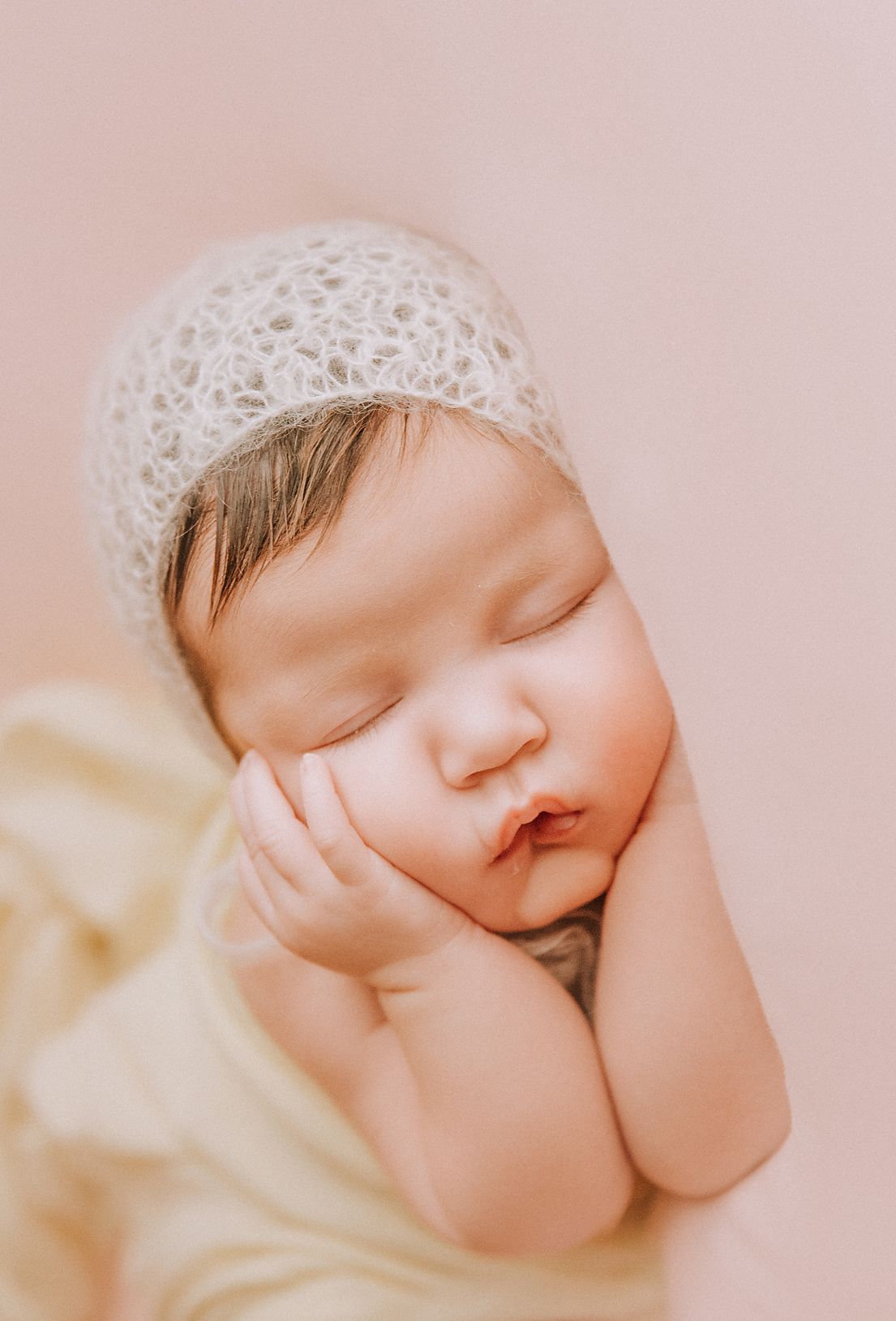 Peaceful newborn baby resting in white lace cap against blush pink backdrop.