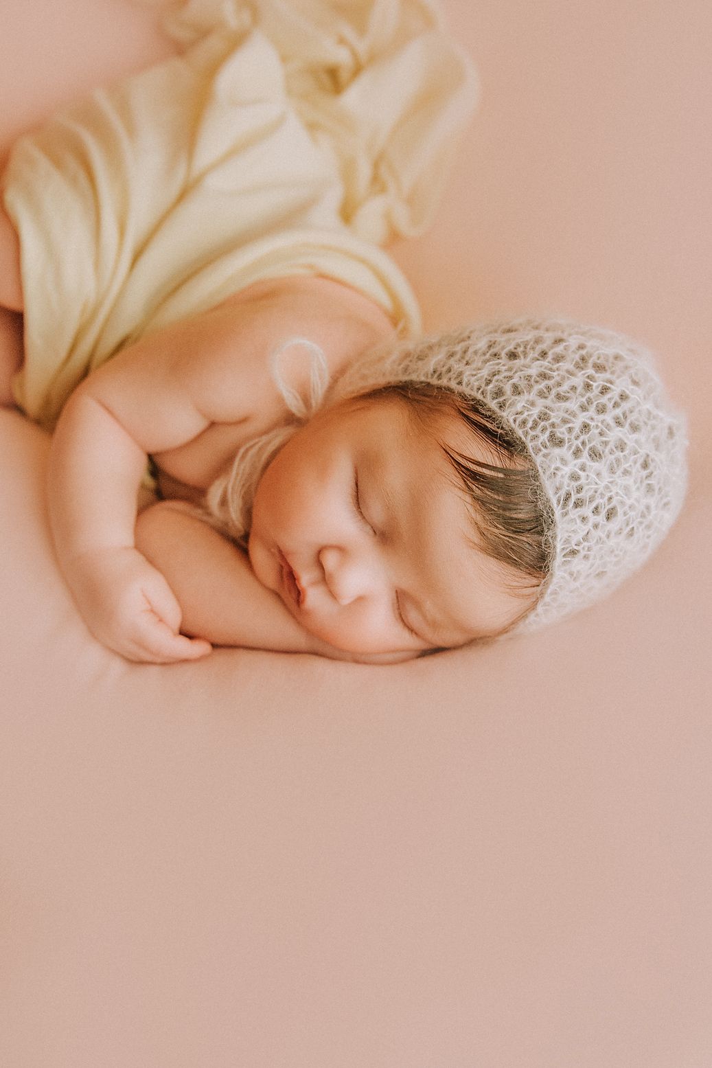 Dreaming newborn baby in cream wrap and lace bonnet on pink background.