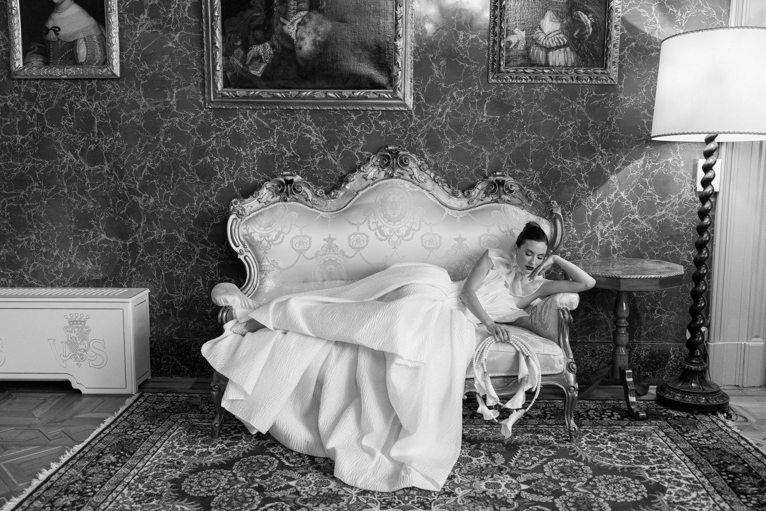 A person in an elegant white dress reclines dramatically on an ornate Victorian sofa in a vintage-style room.
