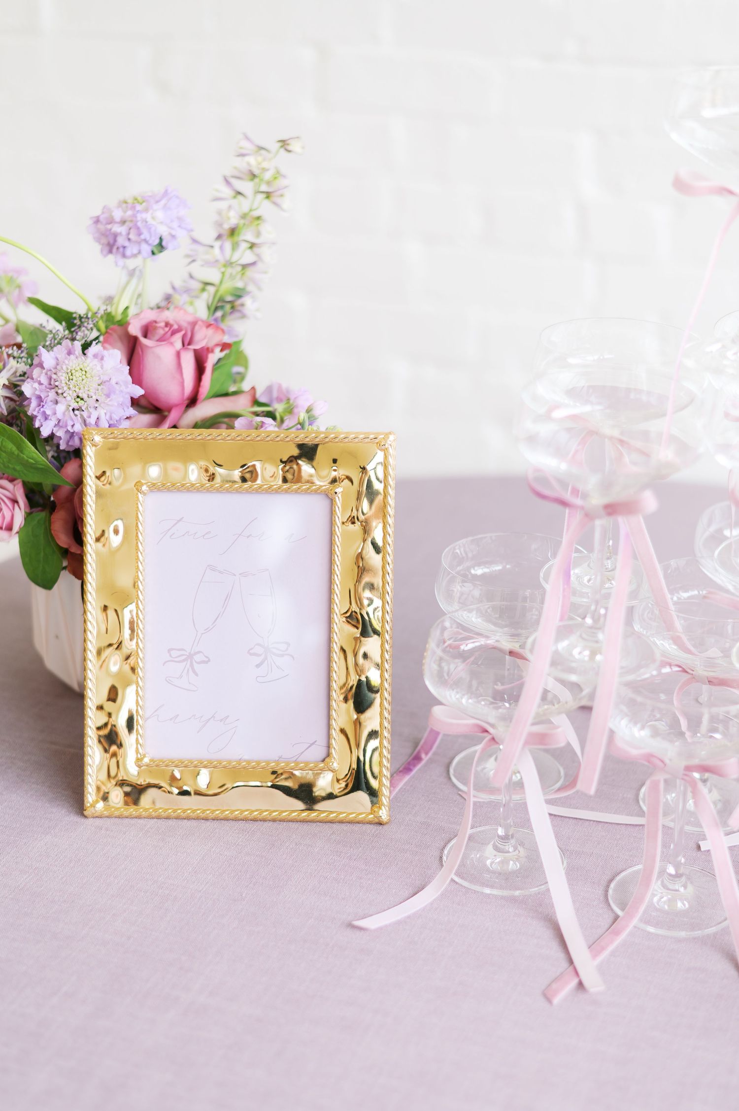 Elegant gold ornate photo frame with pink and purple flowers on a light gray surface with pink ribbons and glass vases.