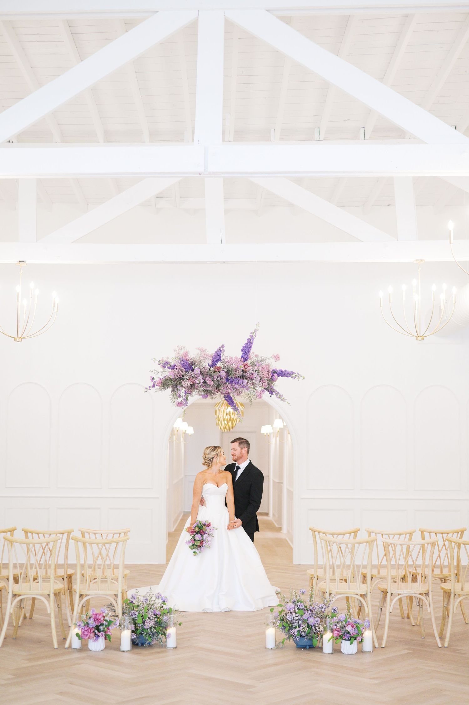 Light and airy wedding venue with wooden chairs, white walls, and purple floral arrangements creating an elegant atmosphere.