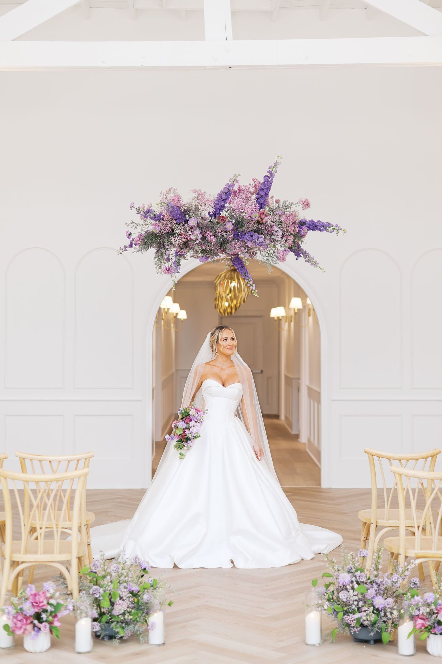 Elegant wedding venue setup with white chairs and purple floral arrangements under arched doorways in a bright interior.