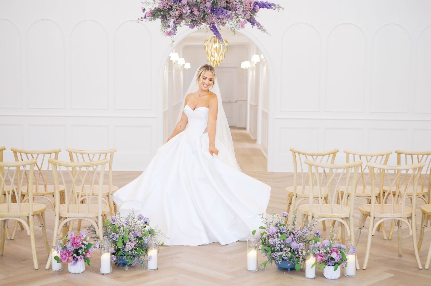 Bride in flowing white gown stands under lavender floral arrangement with natural light streaming through in fayetteville NC
