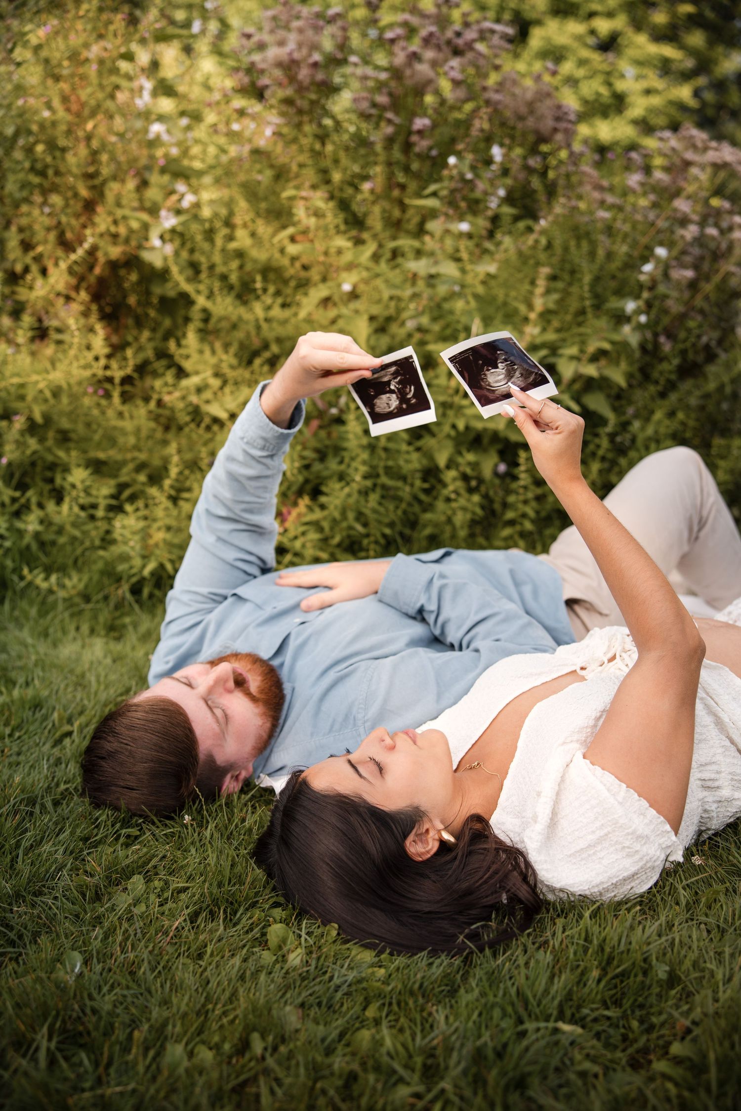 A pregnant couple lying on grass holding ultrasound photos in Amsterdam.