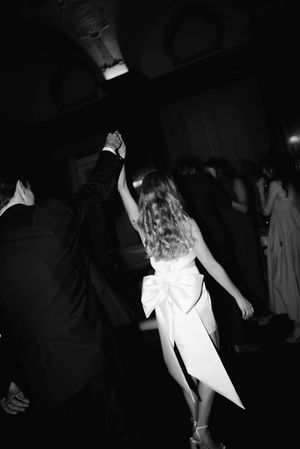 A couple dancing at a formal event, with the woman wearing a white dress with a large bow in black and white photography.