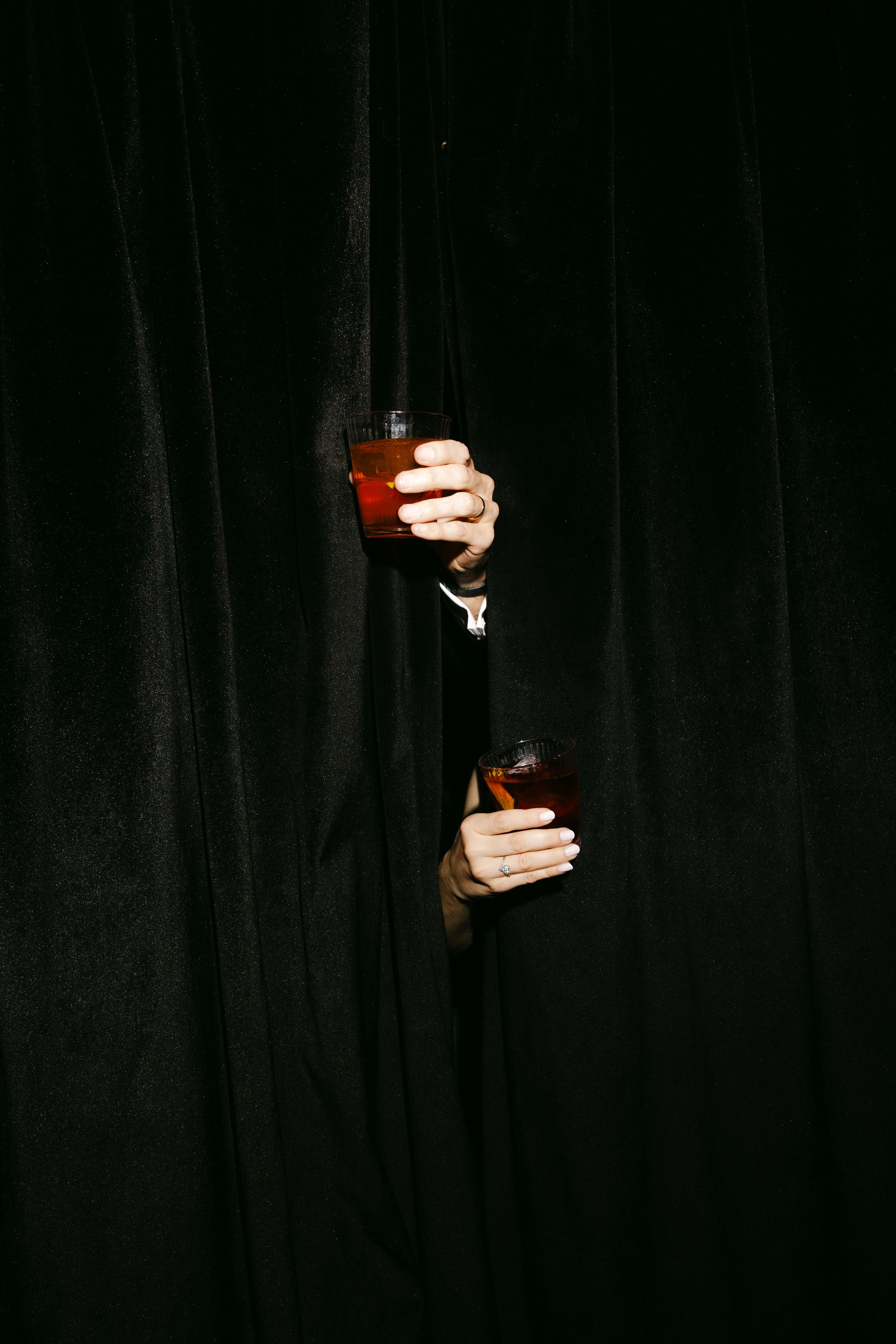 Hands holding drinks against a dark background in dramatic lighting.
