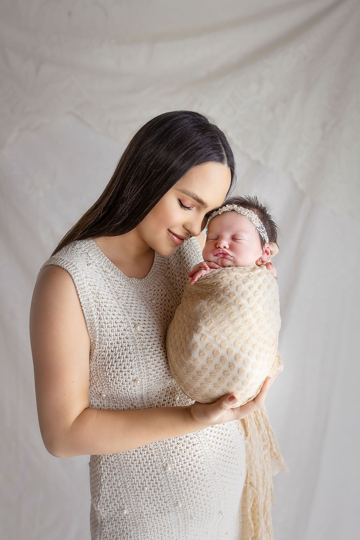mama posa con su bebe ella esta vestida de crema y bebe tambien mama une su cabeza con la bebe en una pose natural