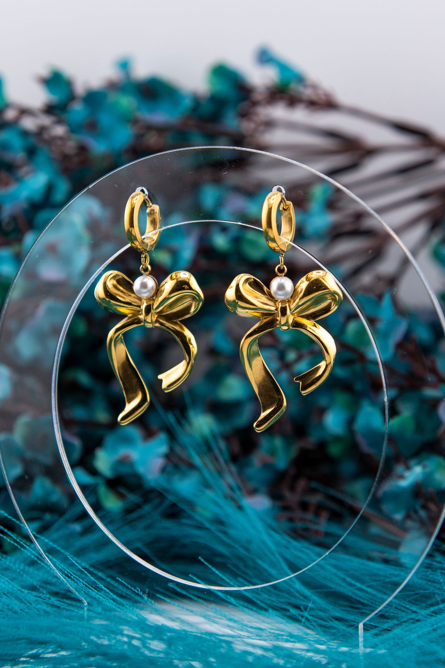 Elegant gold bow earrings displayed on circular glass pieces against a vibrant turquoise backdrop.