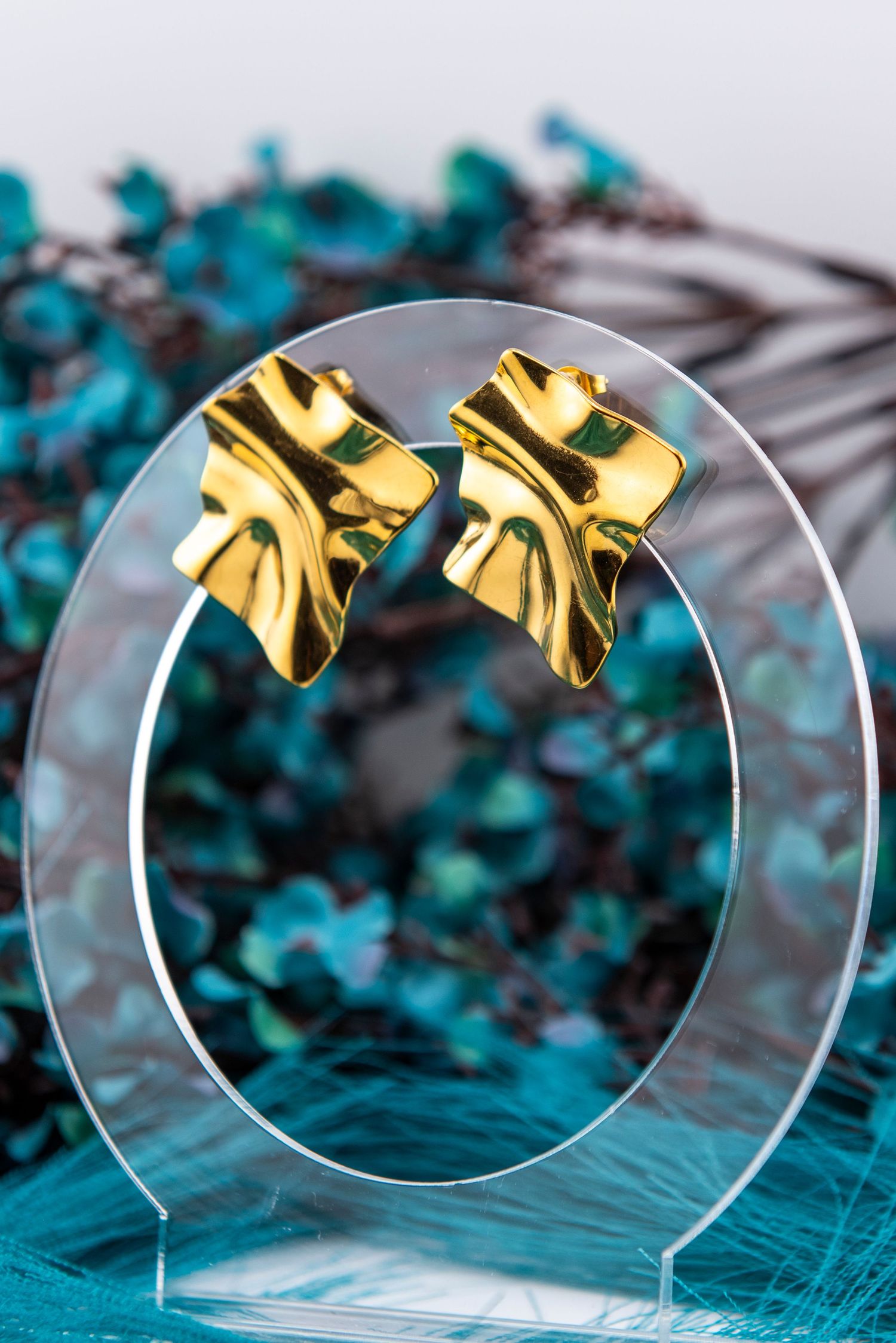 Gold bow-shaped earrings arranged on transparent glass circles with teal floral elements in background.
