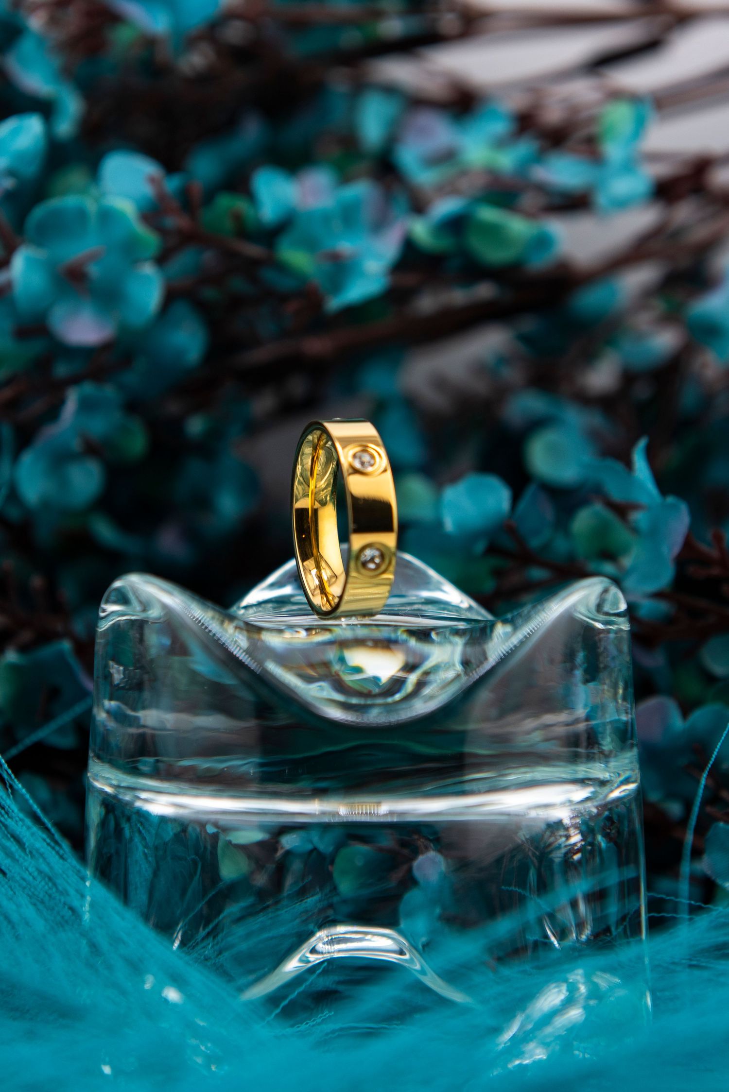 Gold wedding ring displayed on a clear glass surface against a turquoise background with artistic bokeh effect.