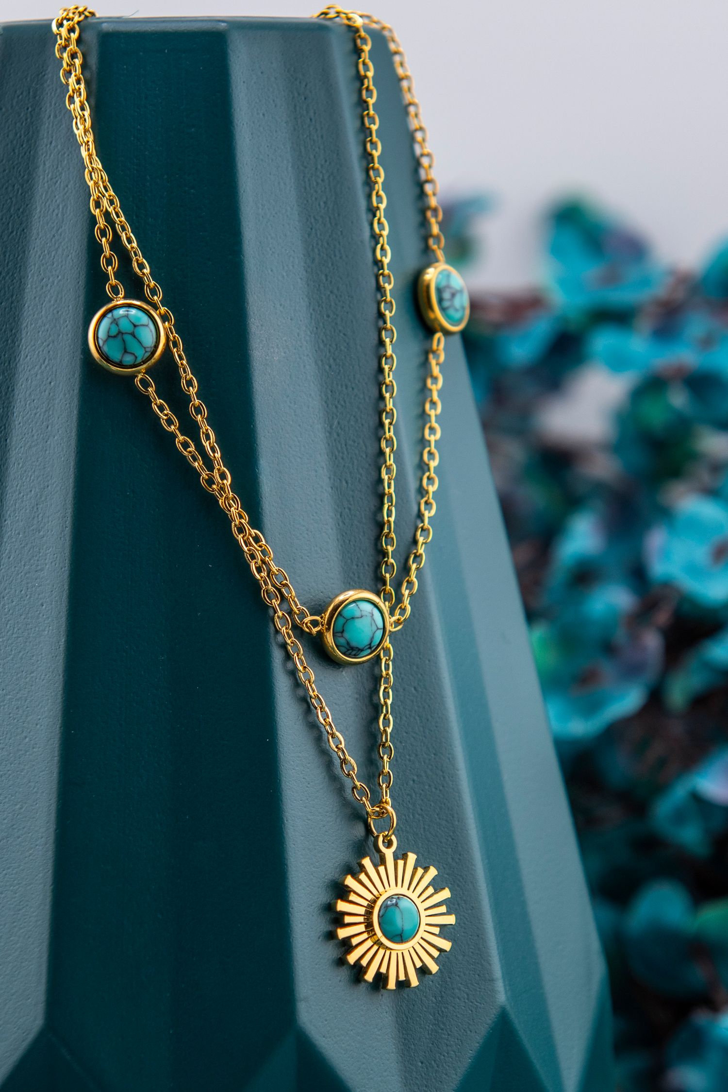 Delicate gold chain necklace with turquoise stones and sunburst pendant displayed against teal background.