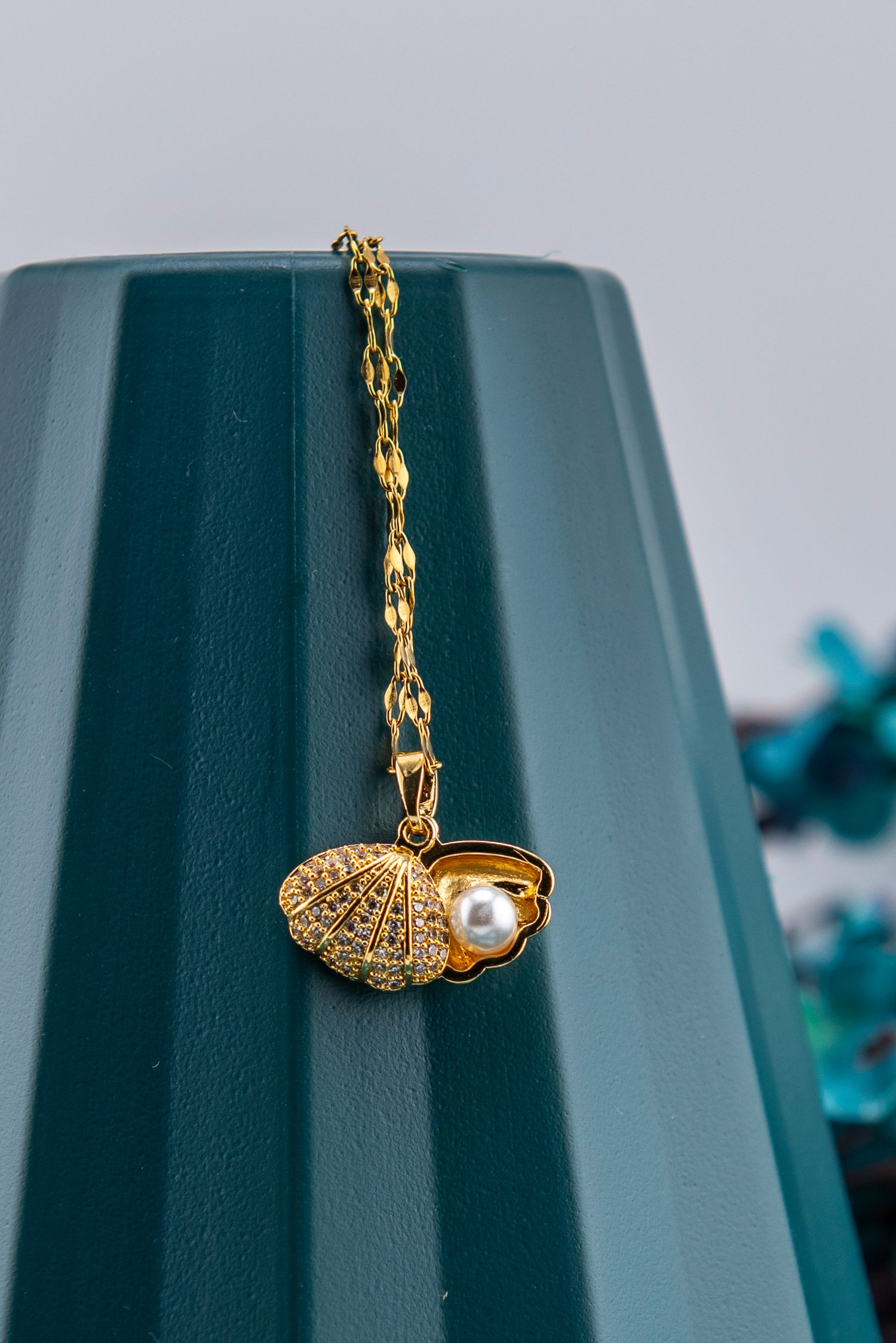 Gold plated necklace with crystal-encrusted shell charm and pearl accent hangs against teal colored background.