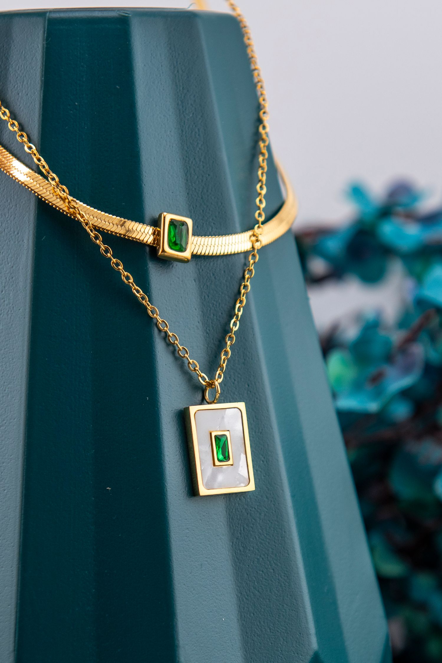 Gold pendant necklace with emerald green gemstone accent displayed against dark teal background.