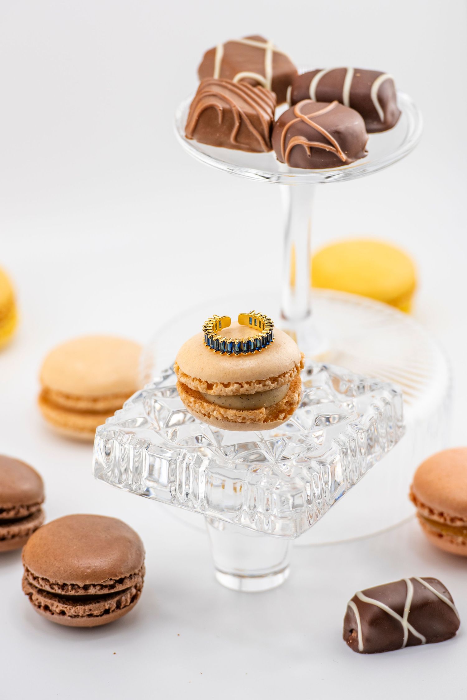 Elegant display of chocolate macarons and truffles on crystal cake stands against a white background.