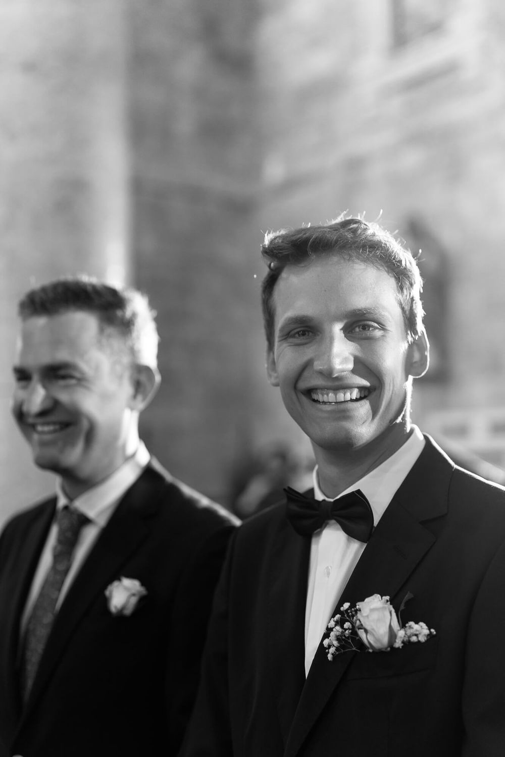 Two grooms smile joyfully in tuxedos with boutonnieres at their wedding ceremony in black and white.