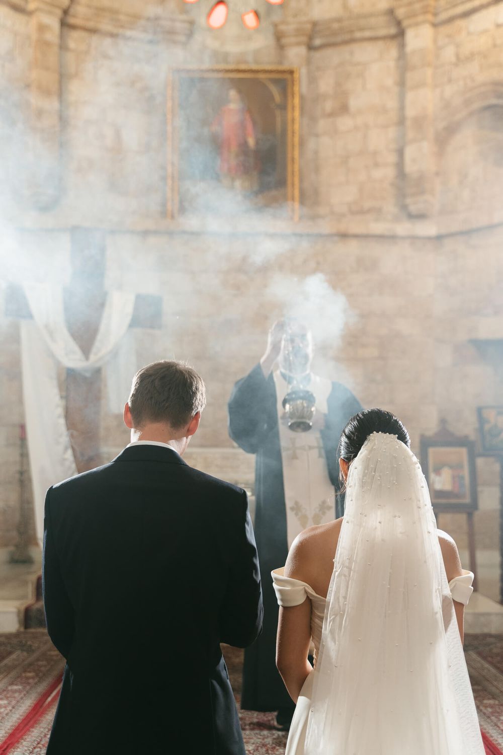 Bride and groom stand before a priest swinging incense during a traditional church wedding ceremony.
