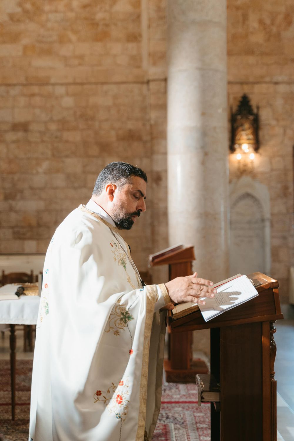 A priest in white vestments reads from a book at a wooden lectern inside a stone church.