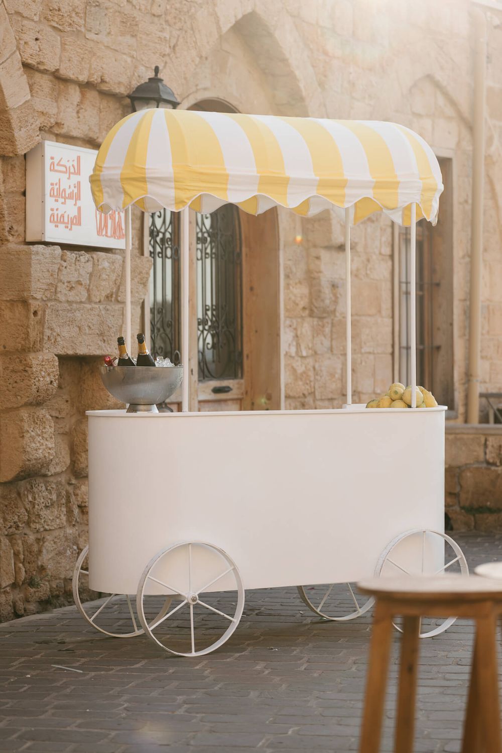White vintage cart with yellow striped awning displaying drinks and fruit on cobblestone street near stone wall.