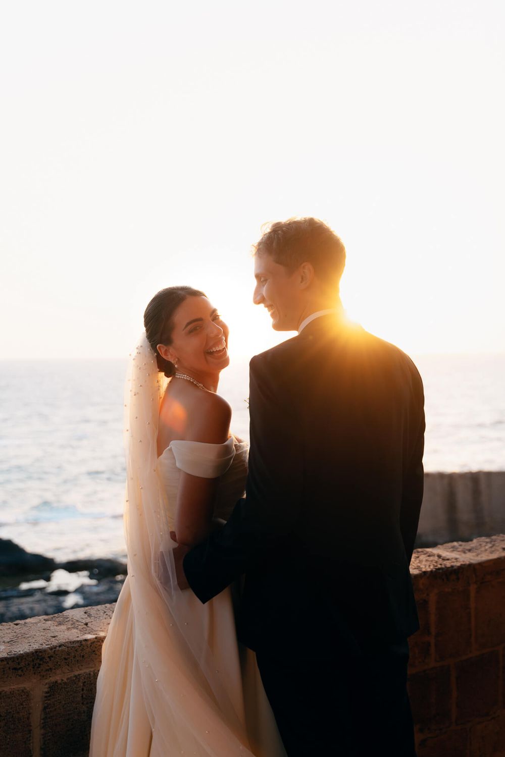 A bride and groom share a joyful moment at sunset by the ocean, with golden light glowing behind them.