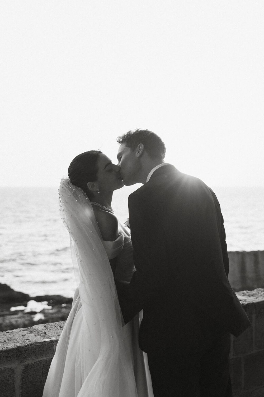 Black and white photo of a bride and groom sharing a kiss by the ocean, with soft backlight and a flowing veil.