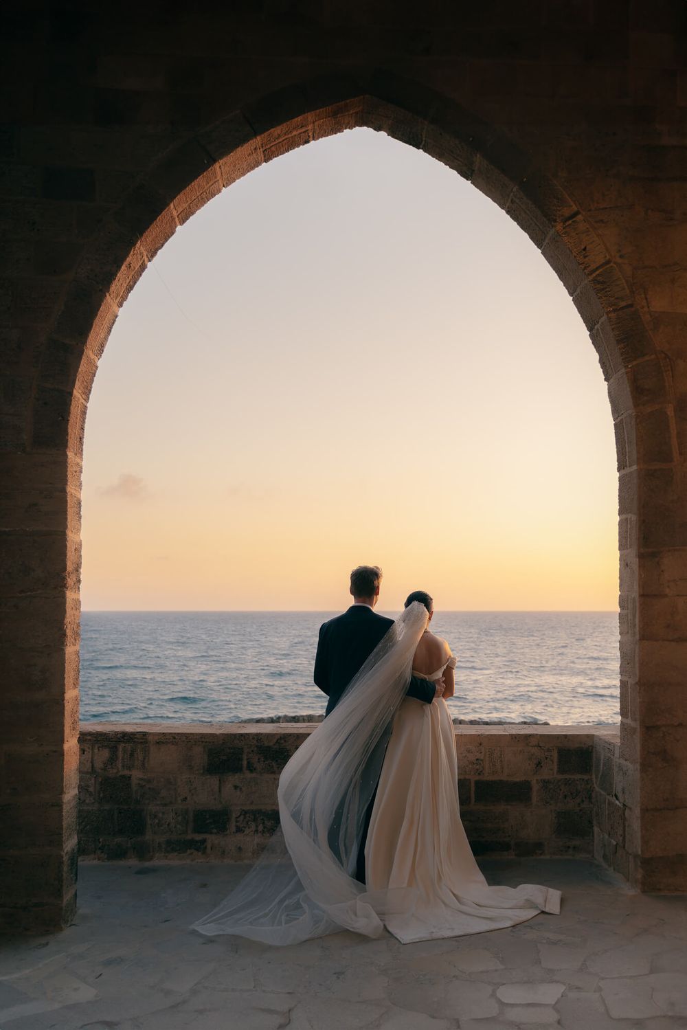 Bride and groom stand under stone arch overlooking ocean sunset at their romantic seaside wedding ceremony.