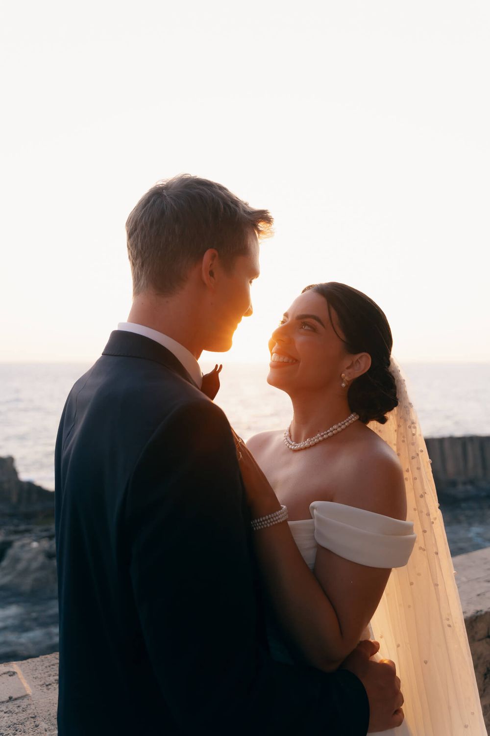 Newlyweds embrace facing each other with golden sunset and ocean backdrop during wedding photo session.